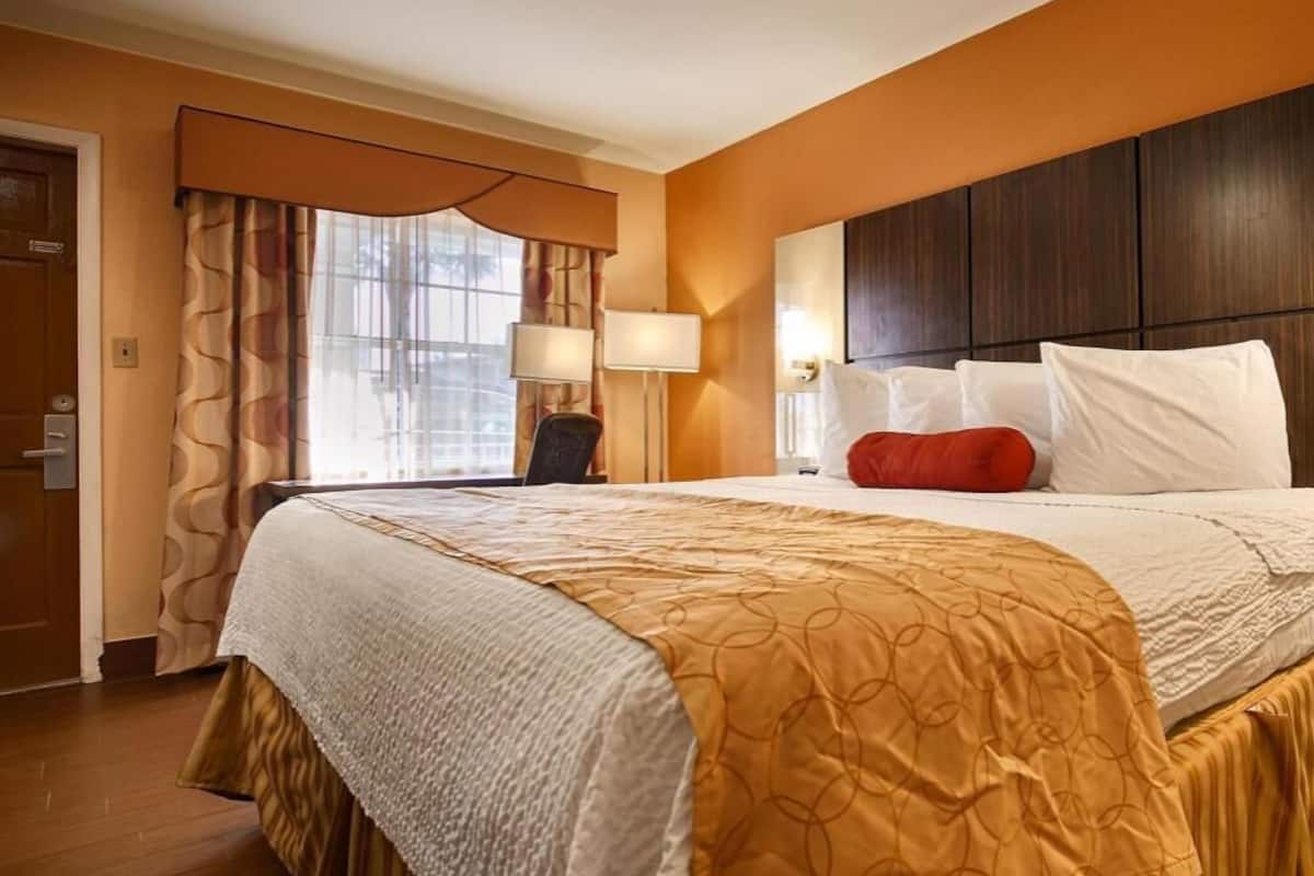 A hotel room with a large bed, a desk, a chair, a window with curtains, and a wall-mounted lamp.