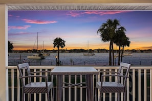 A balcony with wicker chairs and a table, overlooking a waterfront with boats and palm trees during sunset.