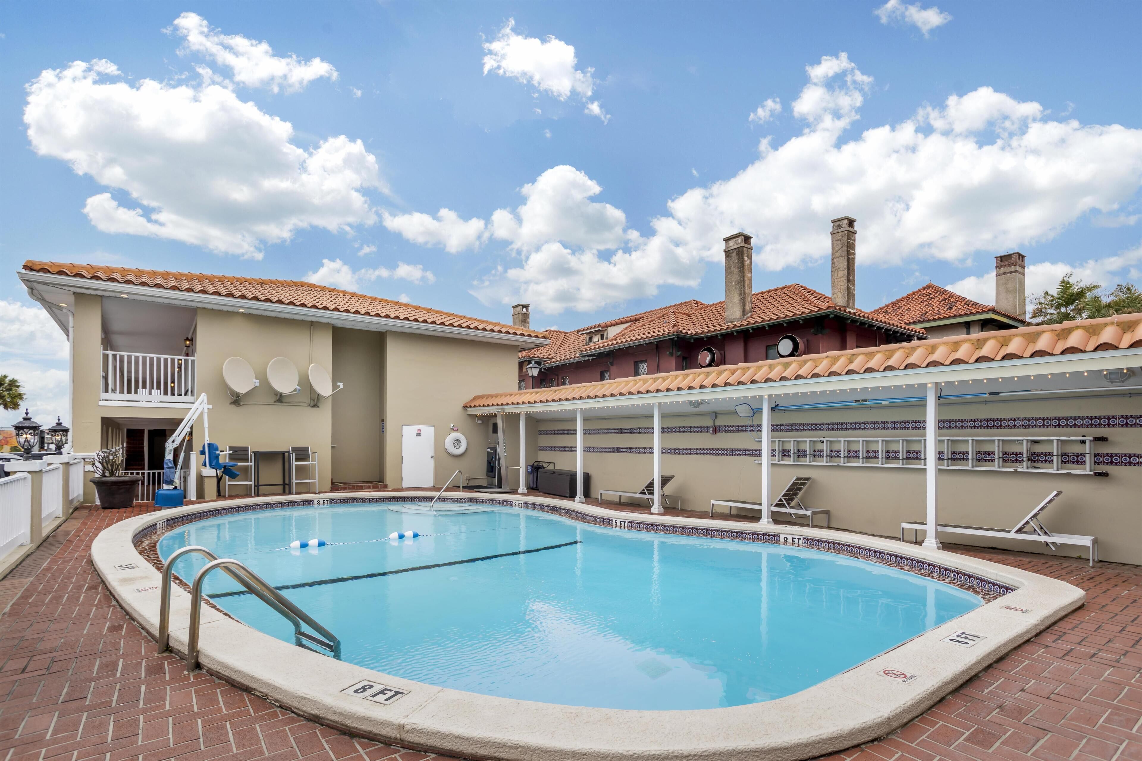 A swimming pool with a tiled deck, a covered area with seating, and a building with a tiled roof and chimneys.