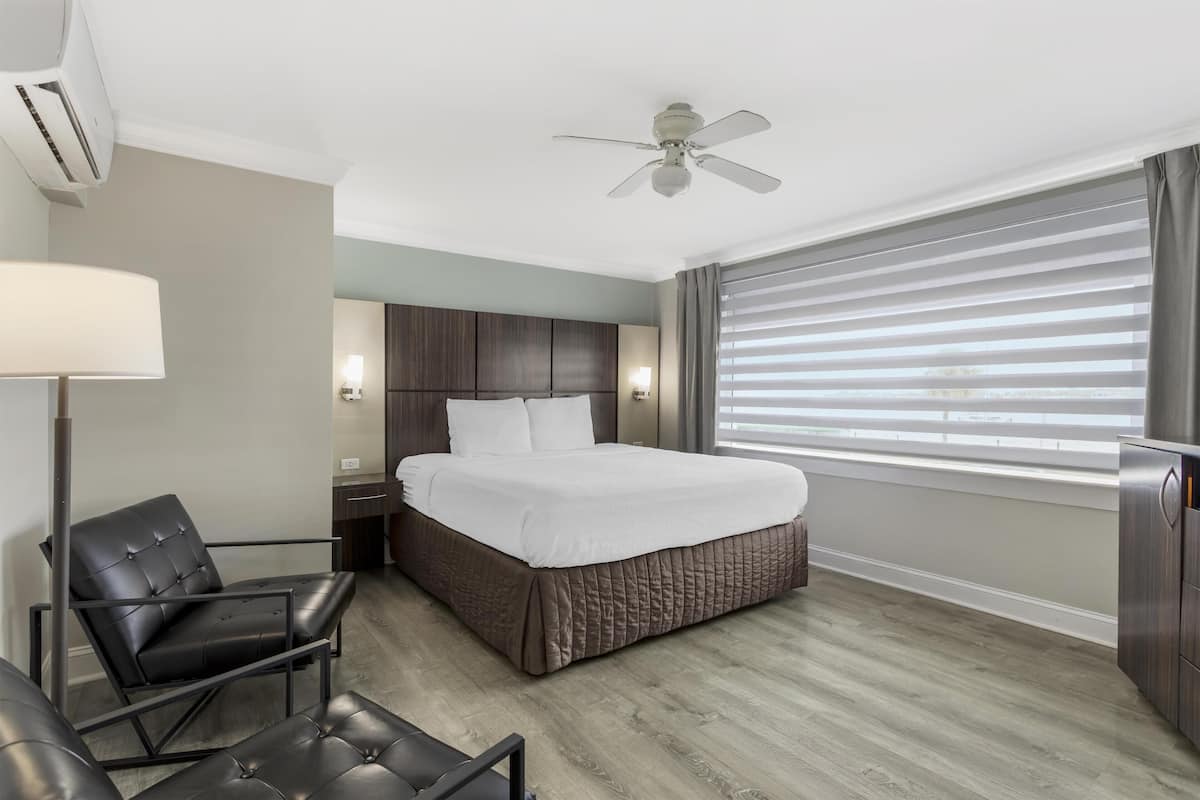 A hotel room with a bed, a chair, a desk, a ceiling fan, and a window with blinds.
