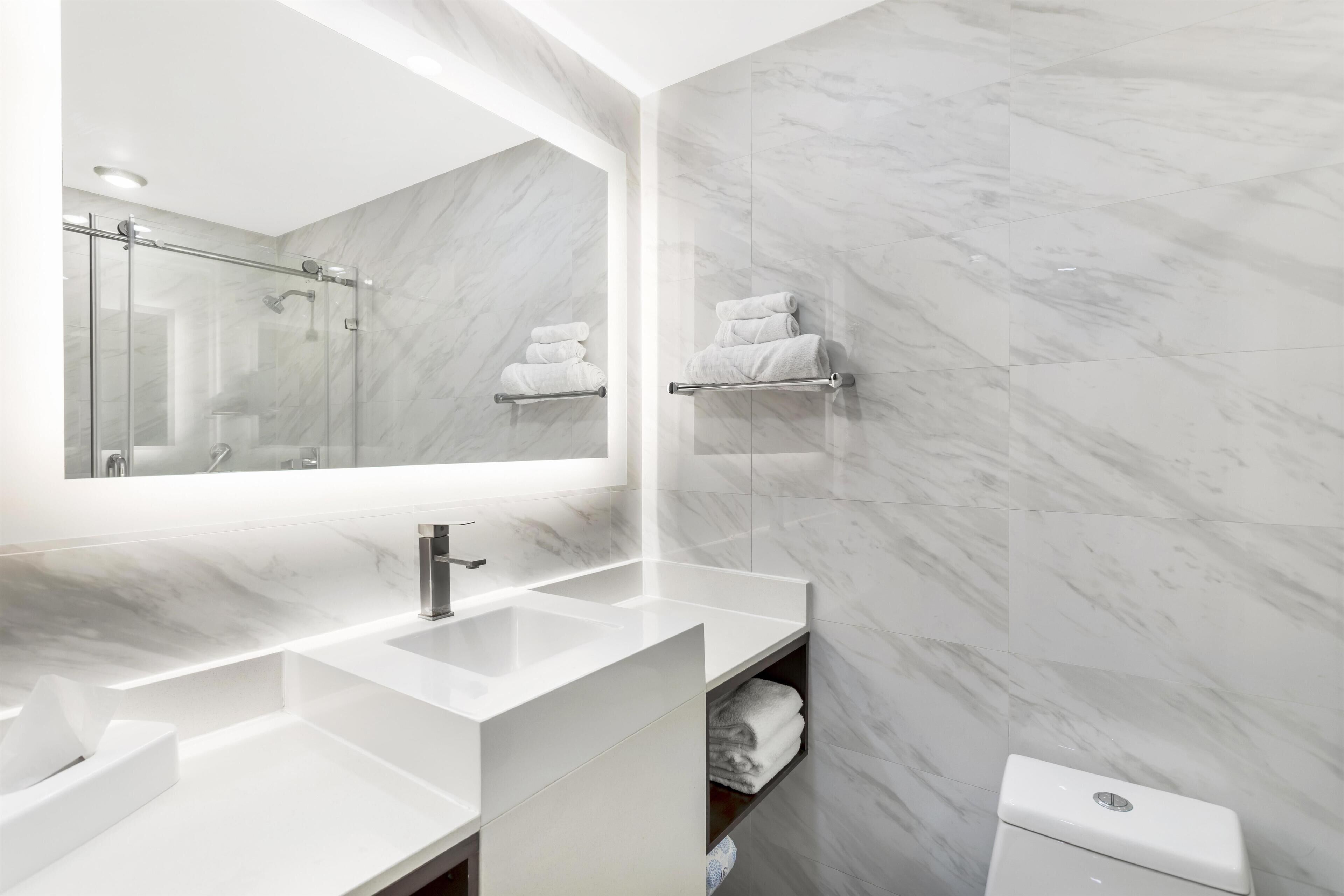 A modern bathroom with a large mirror, two sinks, a shower, and marble walls.