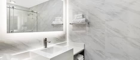 A modern bathroom with a large mirror, two sinks, a shower, and marble walls.