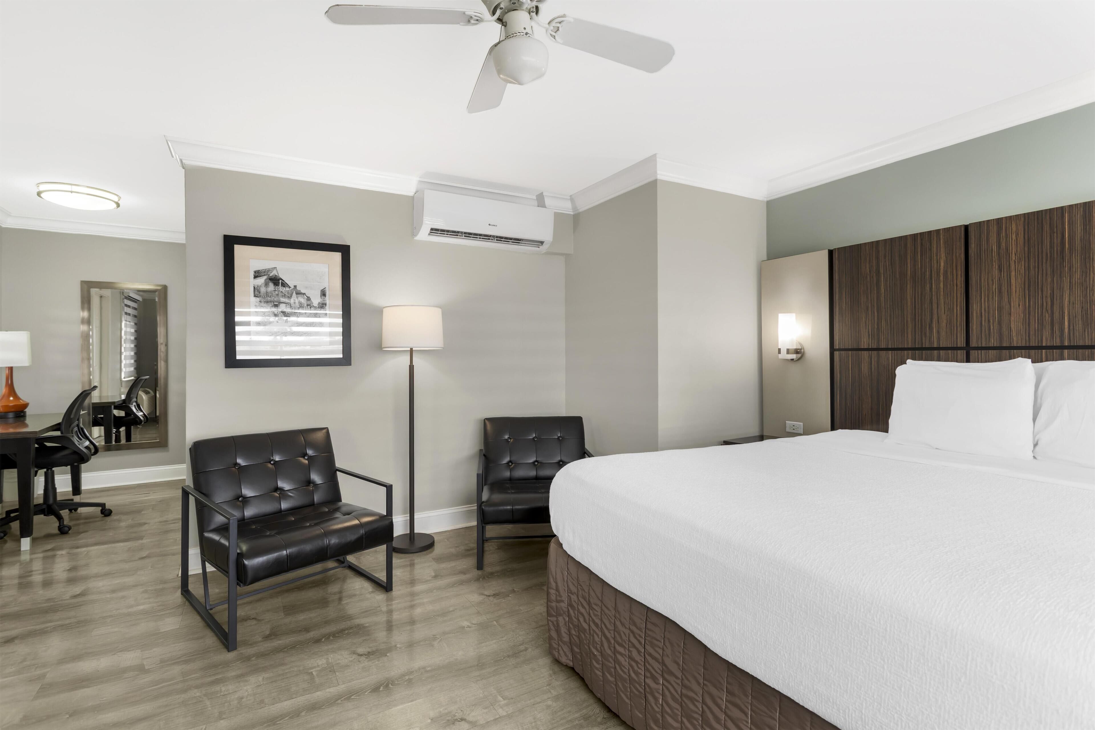 Habitación de hotel con una cama grande, dos sillas, un escritorio con lámpara, equipo de aire acondicionado y ventilador de techo.