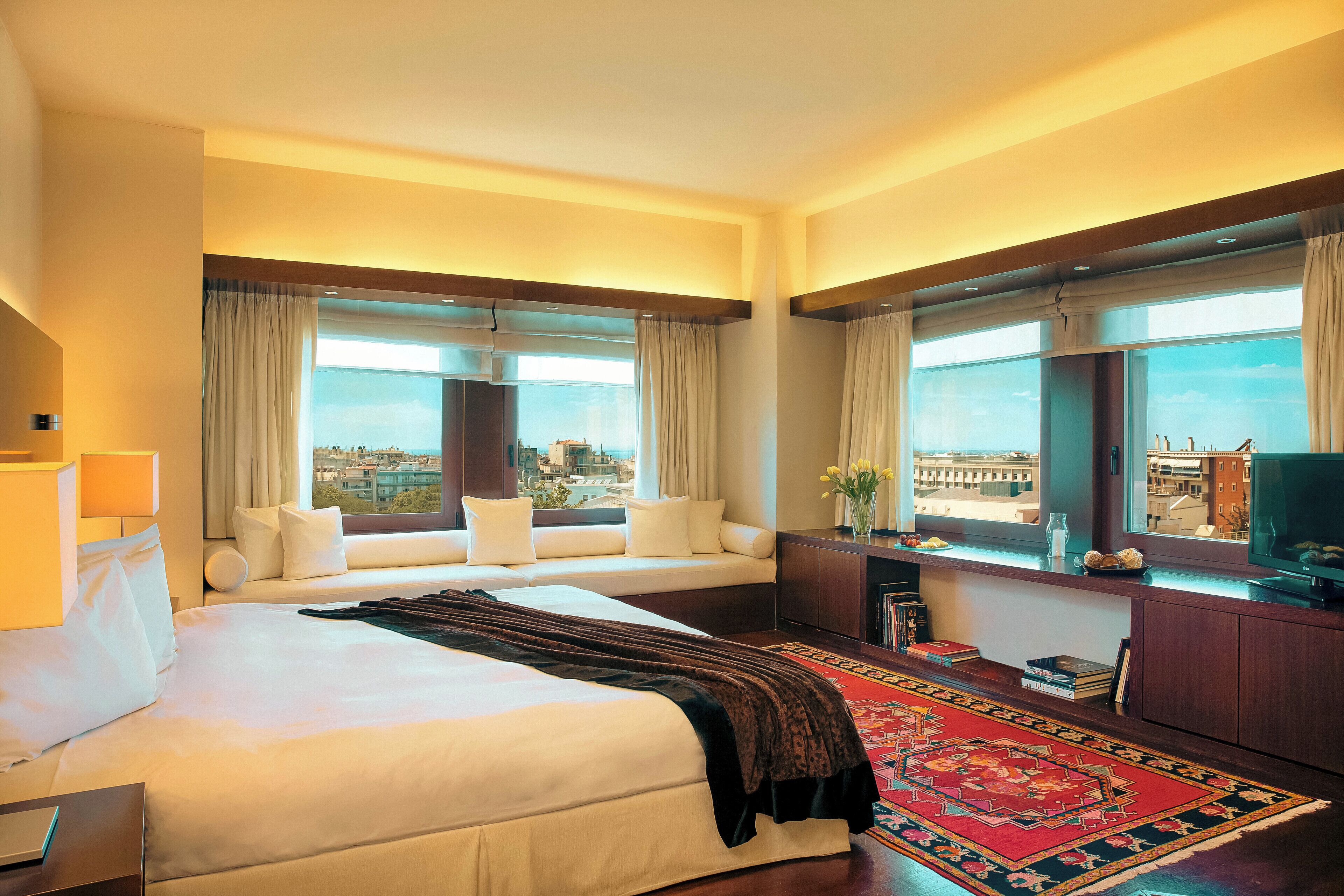 Ein Hotelzimmer mit einem großen Bett, einer Couch, einem Schreibtisch mit Fernseher und Blick auf die Stadt durch große Fenster.