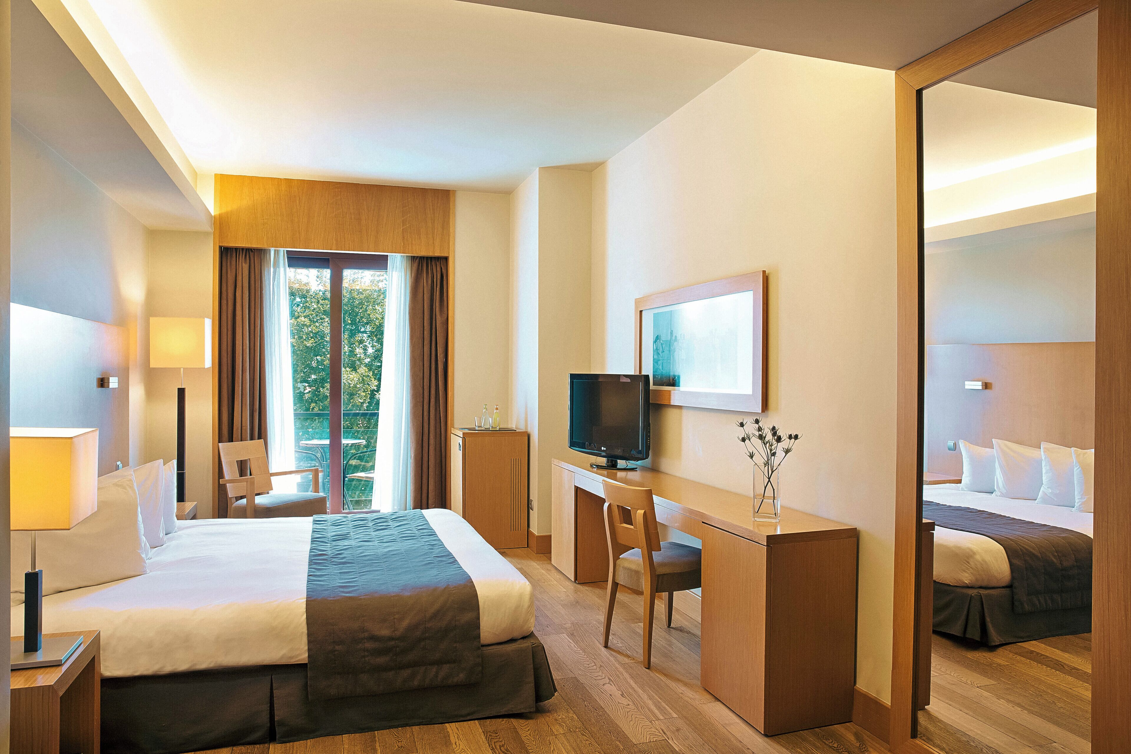 Habitación de hotel con cama, escritorio, silla, televisión y un amplio ventanal con cortinas.