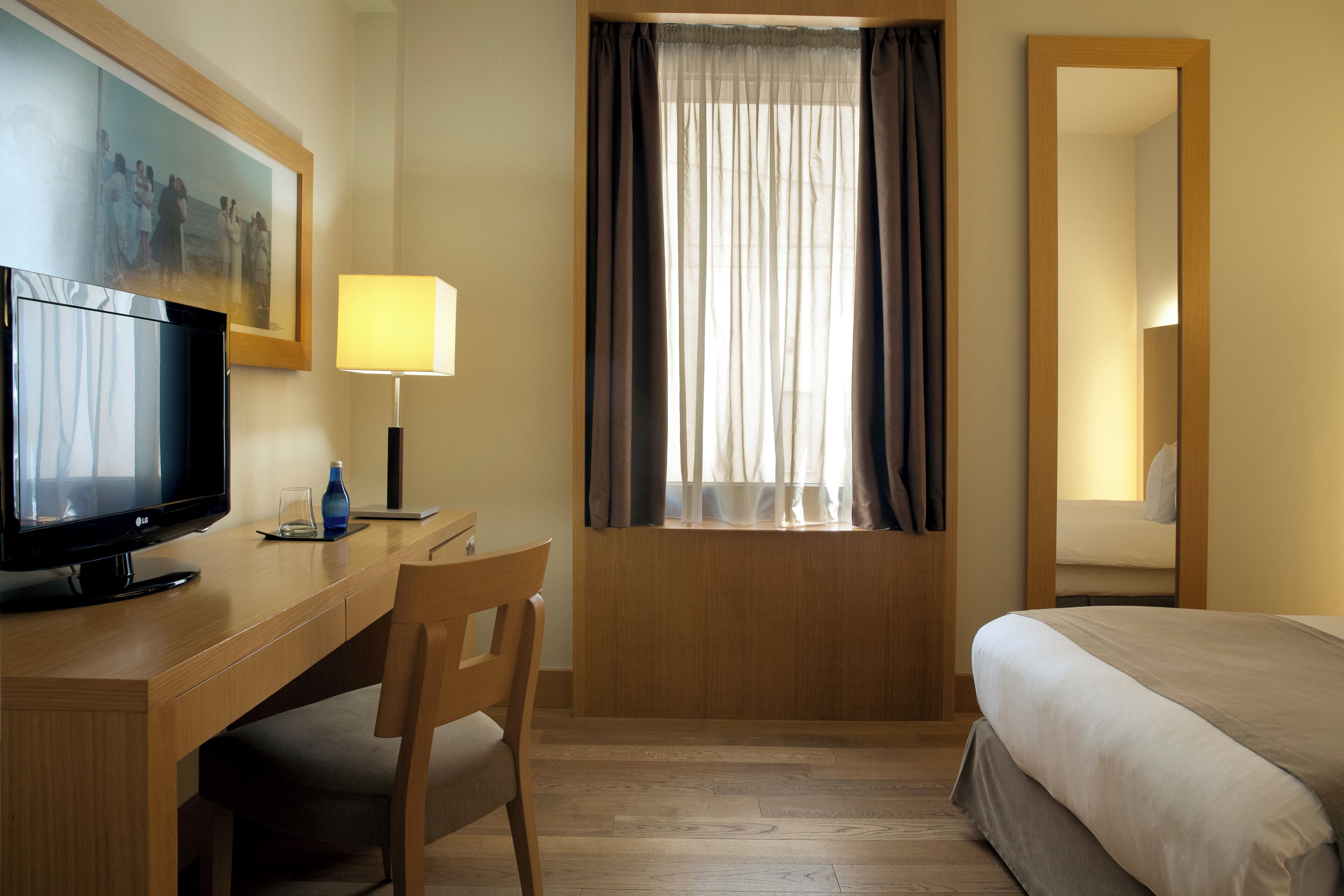 Habitación de hotel con un escritorio de madera, televisión, lámpara, cama y ventana con cortinas.