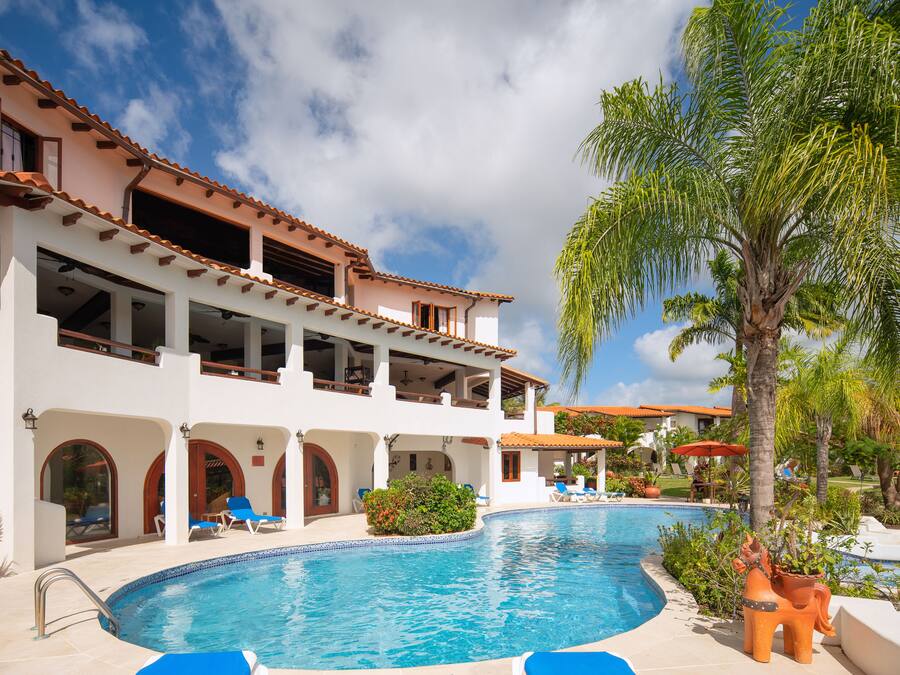 A poolside area with a clear blue pool, white building with red-tiled roof, and palm trees.