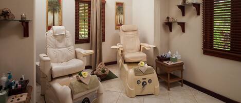 A spa room with two massage chairs, a small table with bottles, and a window with blinds.