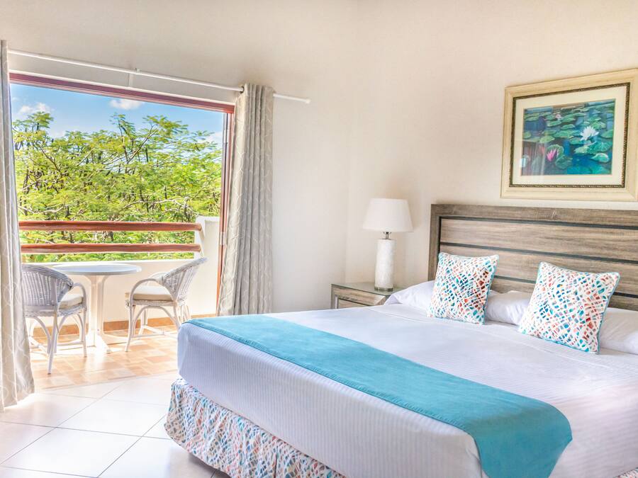 A hotel room with a large bed, a balcony with chairs, and a view of trees.