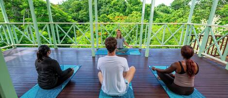 Trois personnes sur des tapis de yoga, dans un espace extérieur abrité, avec vue sur une végétation luxuriante.