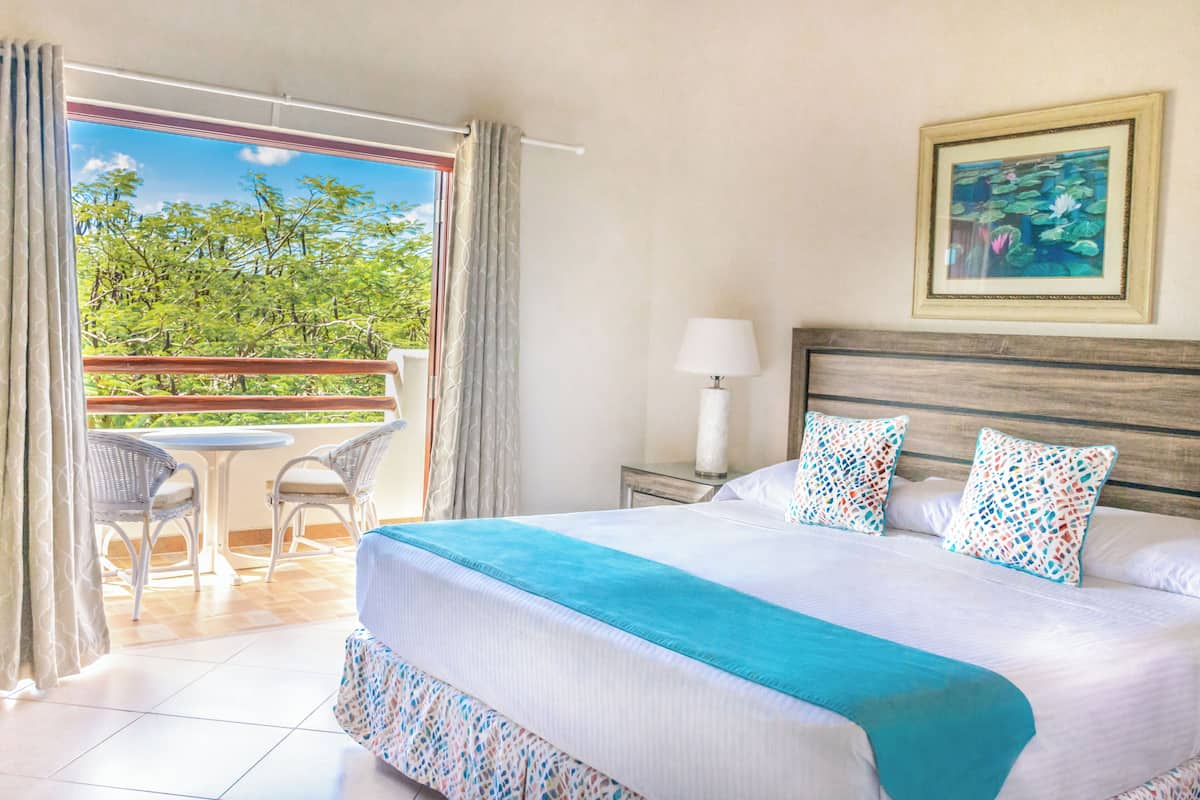 Une chambre d’hôtel avec un lit, une table de chevet, une lampe, un tableau encadré, un balcon avec des chaises et une vue sur les arbres.