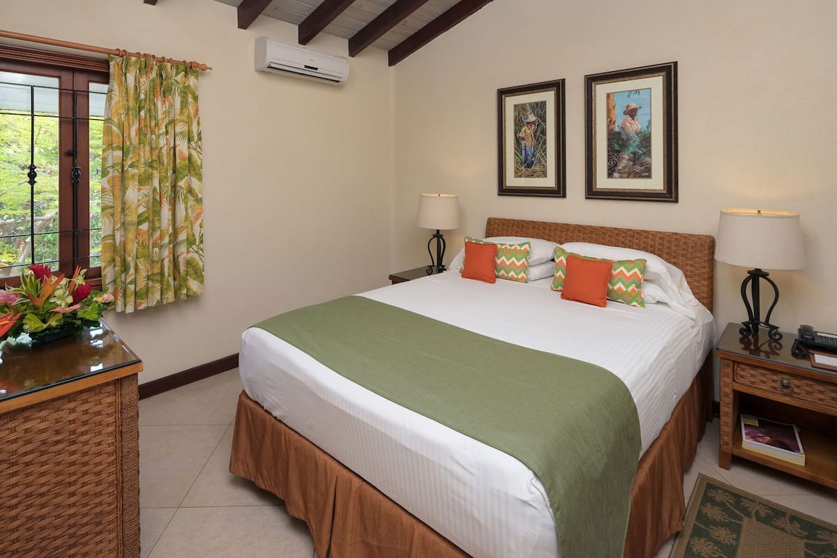 Une chambre d’hôtel comprenant un lit, deux climatiseurs fixés au mur, une fenêtre avec des rideaux, deux tableaux encadrés, une table de chevet avec une lampe et une petite table de chevet avec un téléphone et un livre.