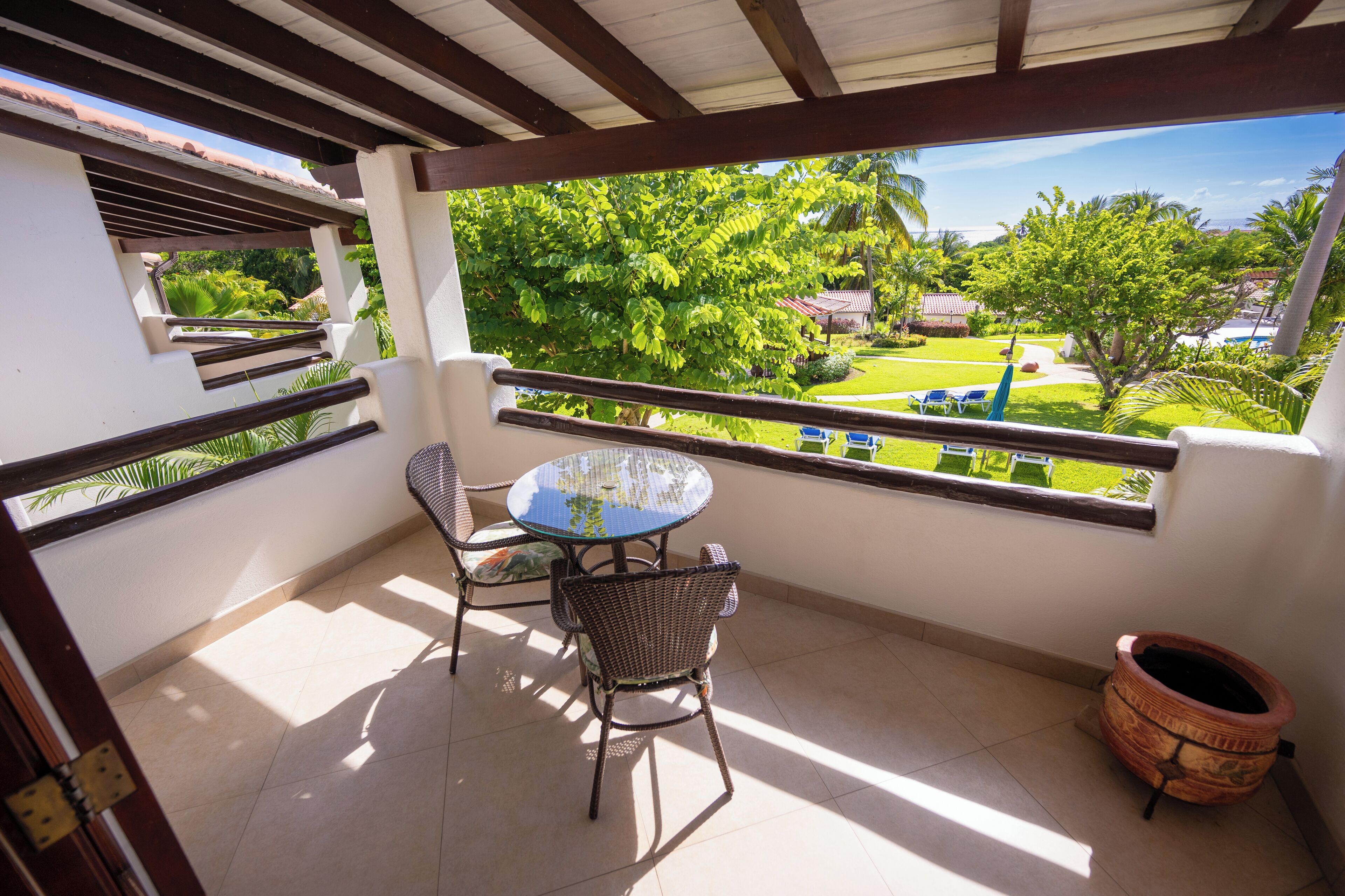 A balcony with a table and chairs, a view of a green garden, and a tiled floor.