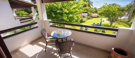 A balcony with a table and chairs, a view of a green garden, and a tiled floor.