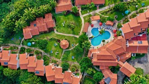 Une vue aérienne d’un complexe hôtelier comprenant plusieurs villas aux toits rouges, une piscine centrale et une végétation luxuriante.