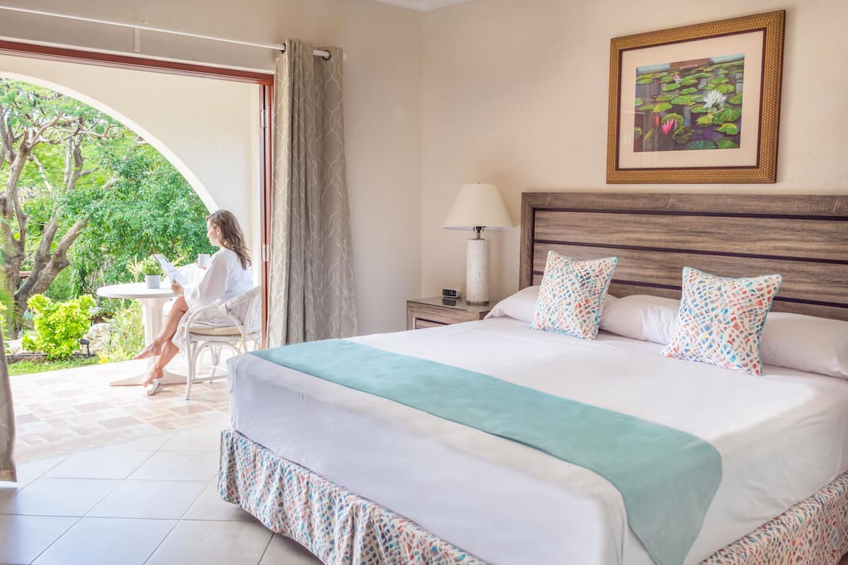 Une chambre d’hôtel avec un grand lit, une table de chevet avec une lampe, un tableau encadré au mur et une vue sur un jardin par une porte ouverte.