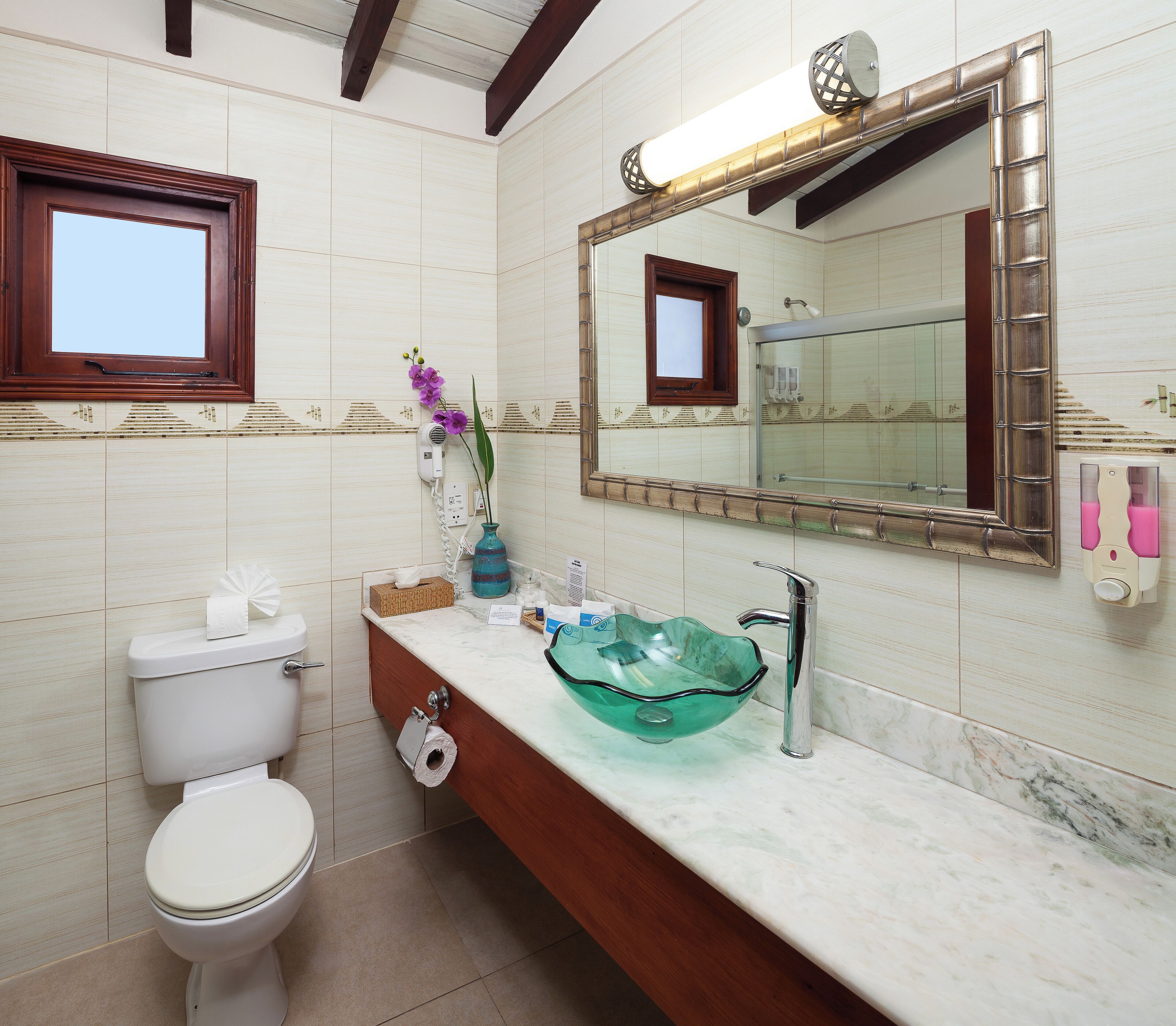A bathroom with a toilet, sink with a glass bowl, a mirror, and a window.