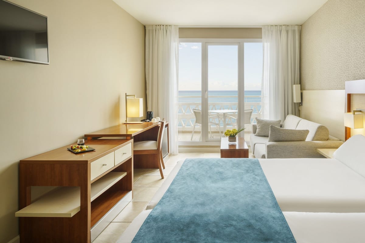 A hotel room with a bed, a desk, a chair, a small table with a lamp, and a view of the sea through a large window.