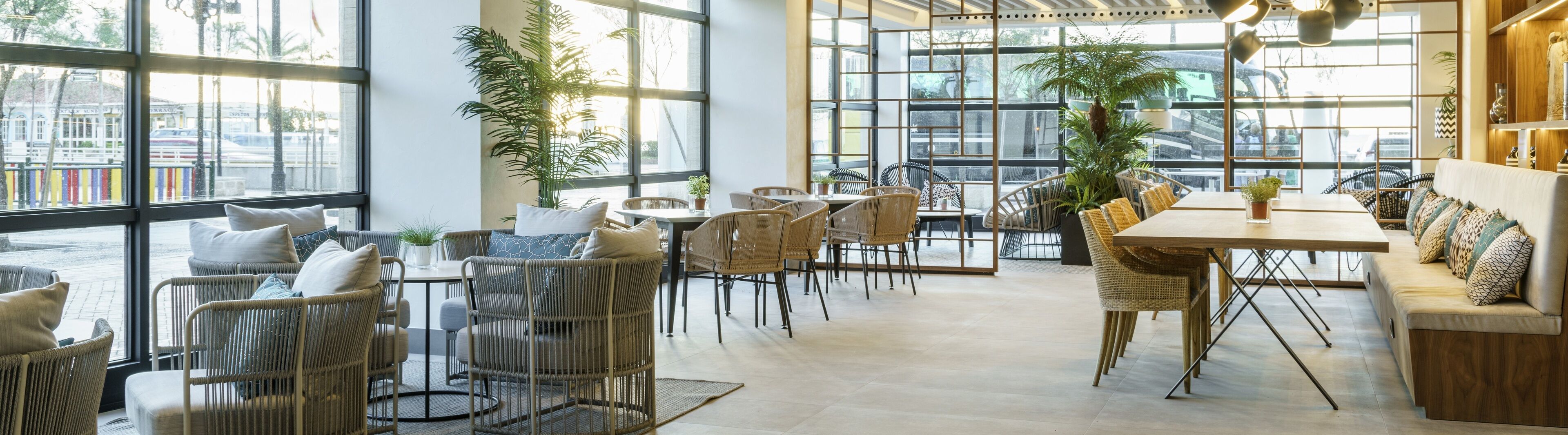 A spacious cafe with wicker furniture, large windows, and indoor plants.