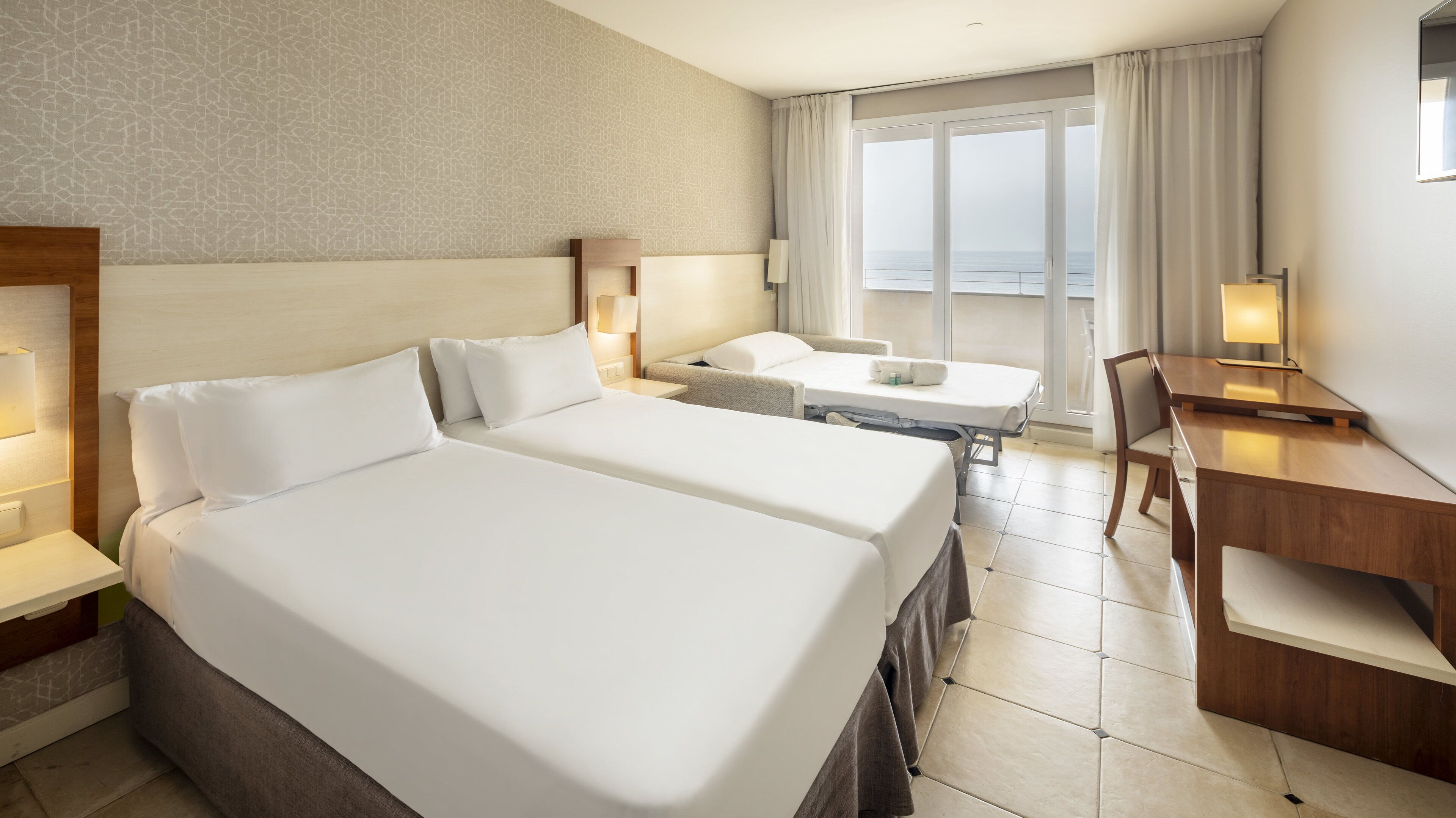 Ein Hotelzimmer mit zwei Betten, einem Schreibtisch und einem Sessel. Ein großes Fenster bietet Meerblick.