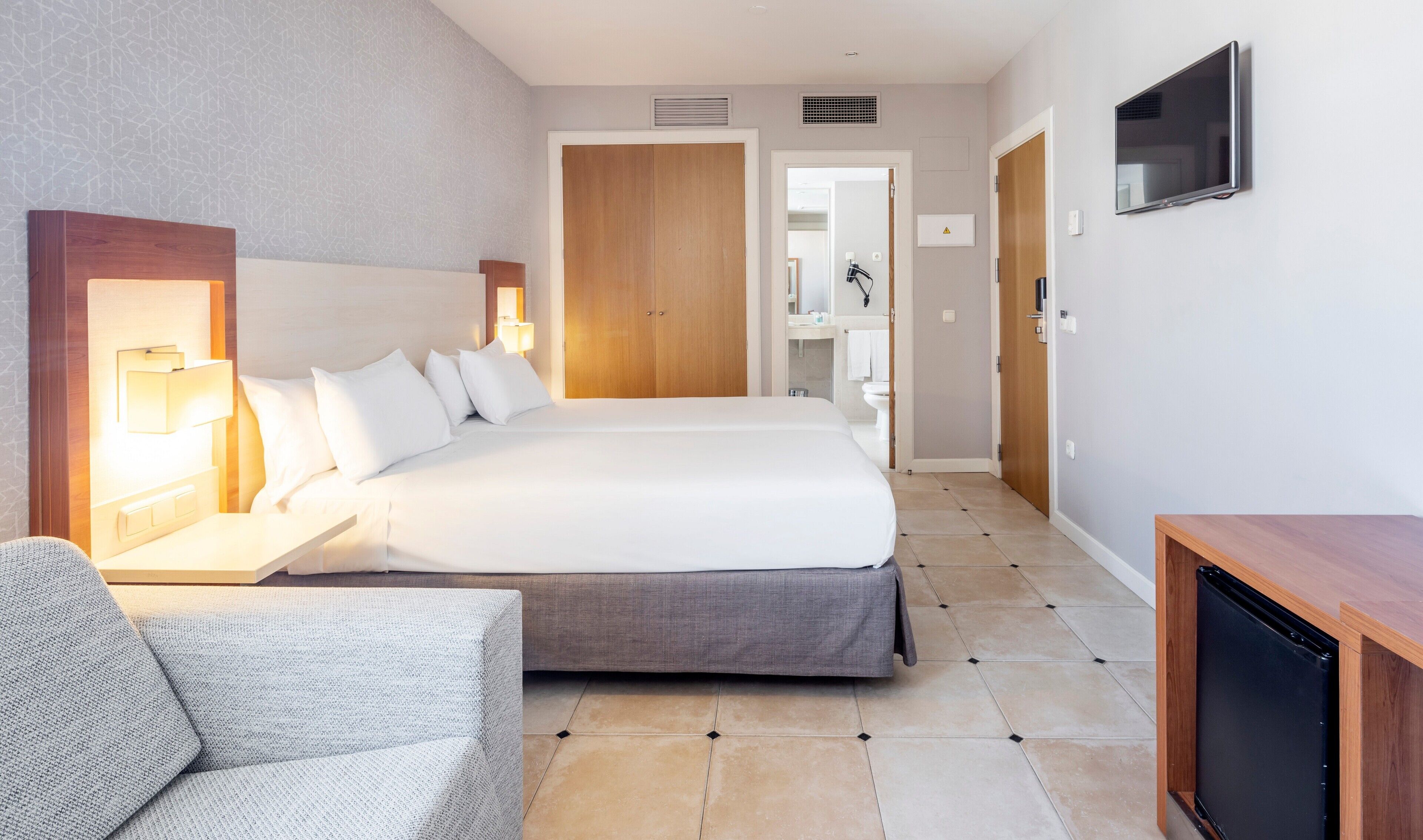 Una camera d'albergo con un letto grande, comodini con lampade, una TV a muro e una piccola scrivania con un computer.
