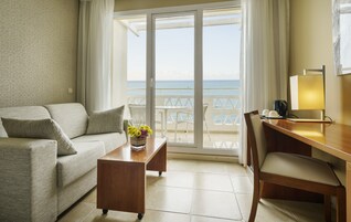 A hotel room with a sofa, a small wooden table with a vase of flowers, a desk with a lamp, and a view of the sea through a sliding glass door.