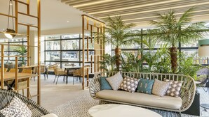 Un espacio interior moderno con sofá, sillas y mesas. Amplias ventanas ofrecen vistas a la calle.