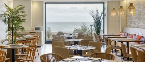 A restaurant with tables and chairs arranged for dining, a large window offering a view of the sea, and a brick wall on one side.