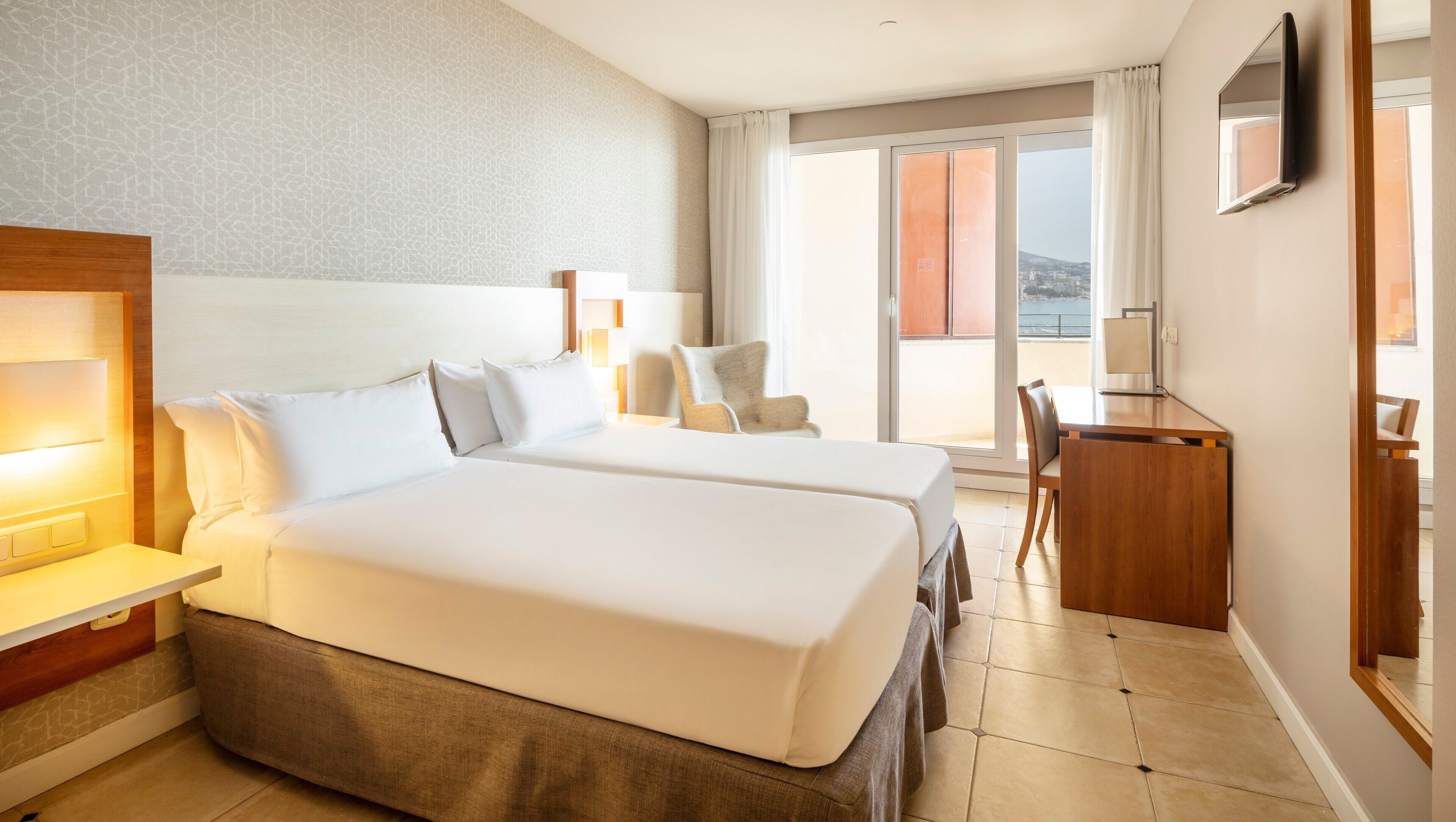 A hotel room with two beds, a desk, a chair, a TV, and a balcony with a view.
