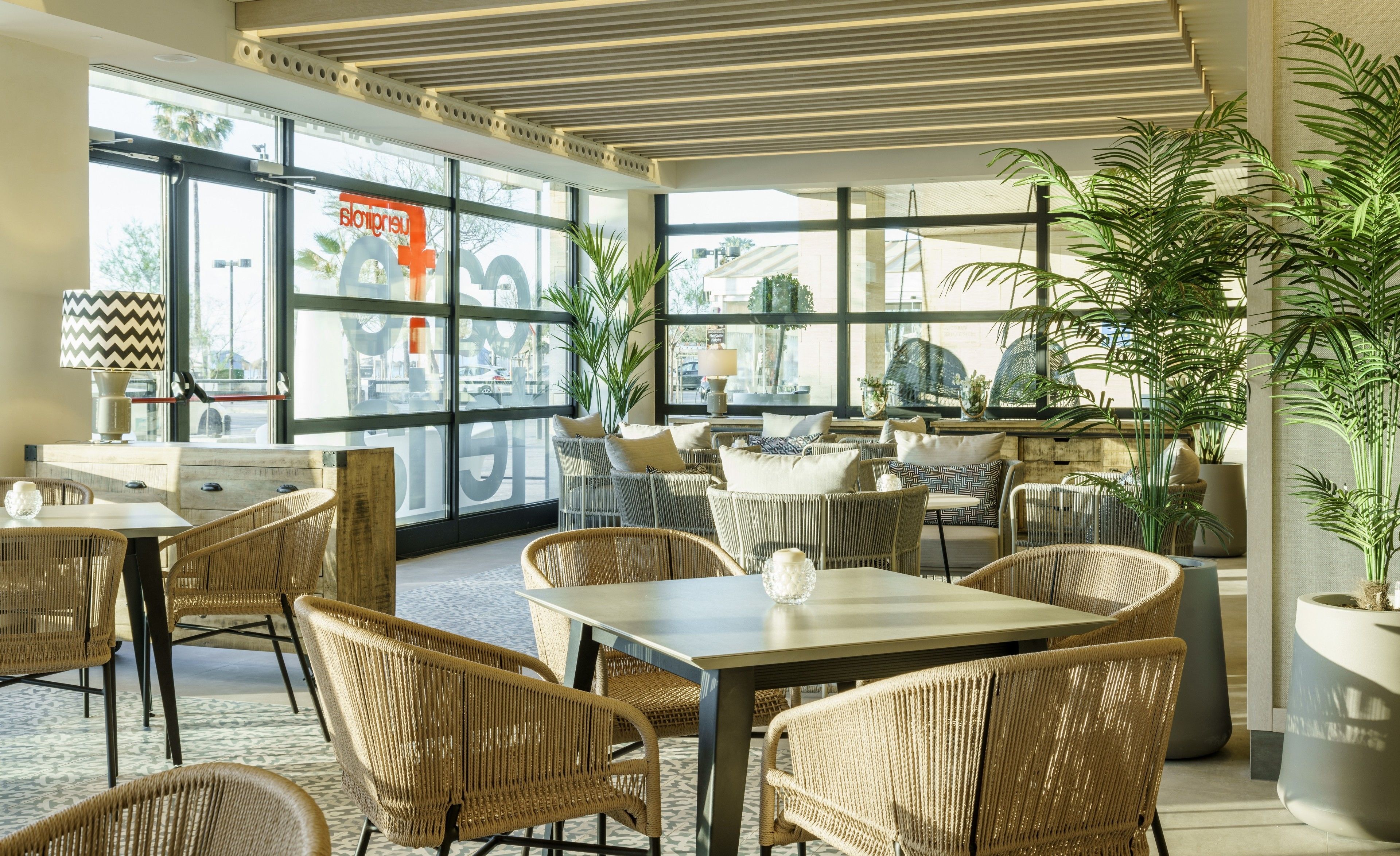 A modern restaurant with wicker furniture, large windows, and indoor plants.