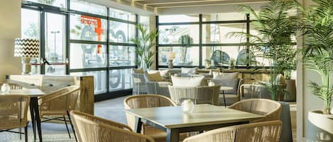 A modern restaurant with wicker furniture, large windows, and indoor plants.