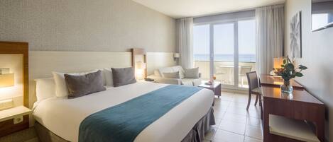 A hotel room with a large bed, a desk, a chair, and a balcony with a view of the sea.