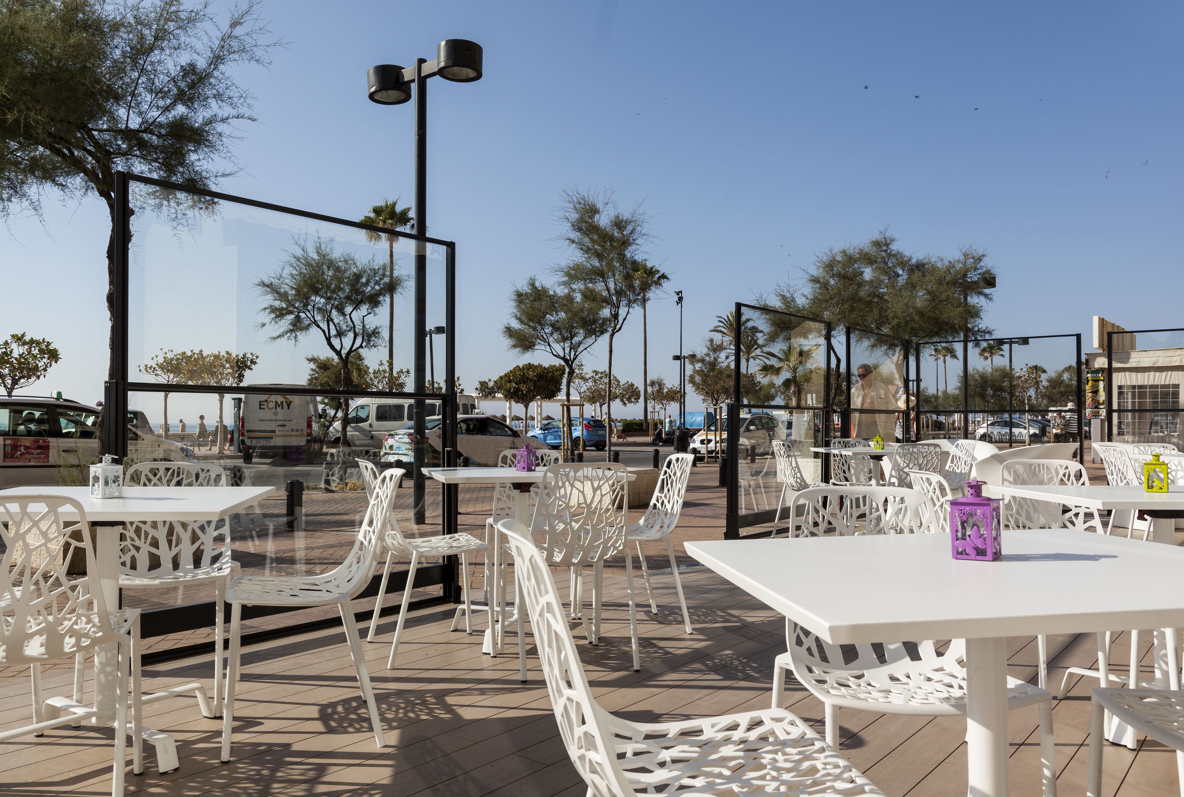An outdoor cafe with white metal tables and chairs, surrounded by trees and a clear sky.