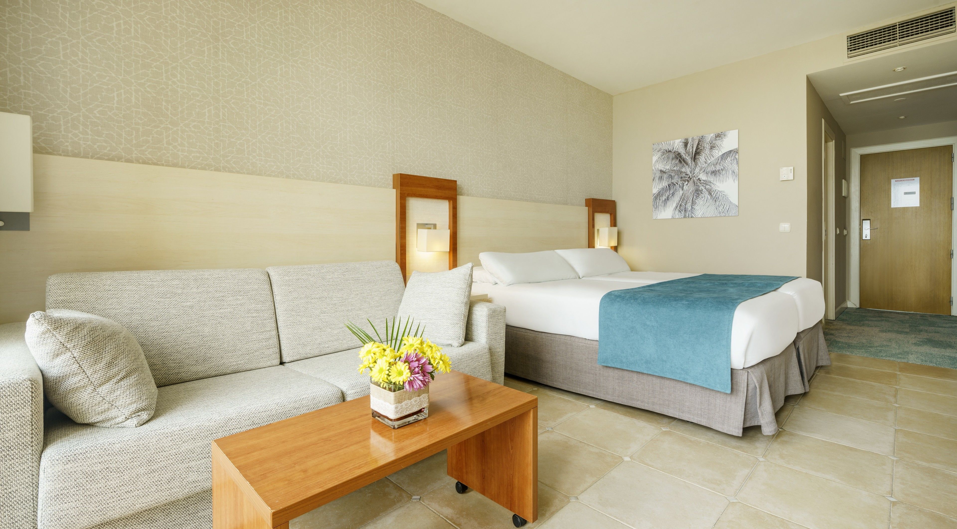 Habitació d'hotel amb un sofà, un llit amb llençol blau, una taula de centre de fusta amb un ram de flors i una làmpada penjada a la paret.