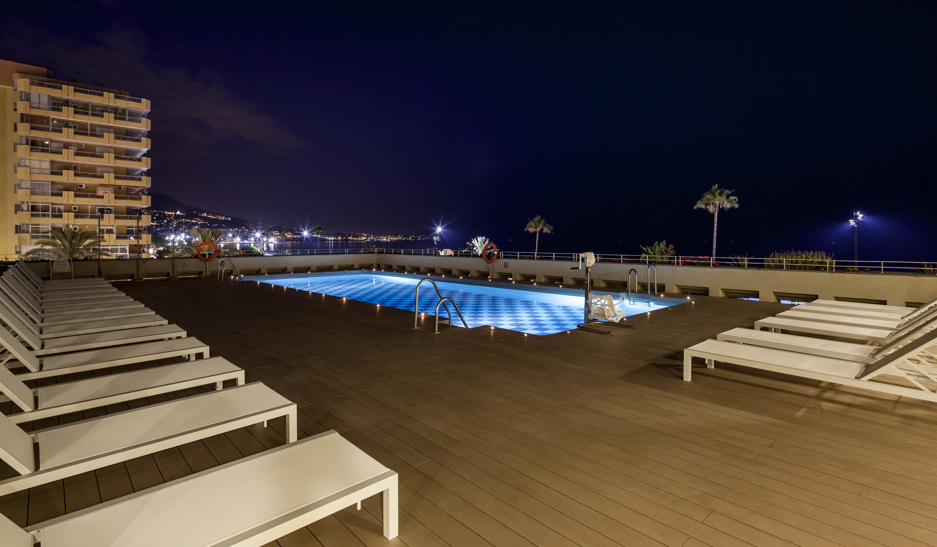 Una piscina en la azotea con sillones de descanso y vista nocturna de la ciudad.