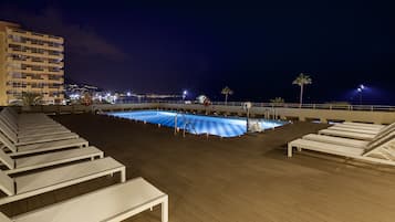 Una piscina en la azotea con sillones de descanso y vista nocturna de la ciudad.