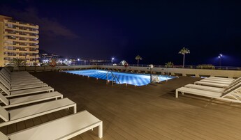 Una piscina en la azotea con sillones de descanso y vista nocturna de la ciudad.