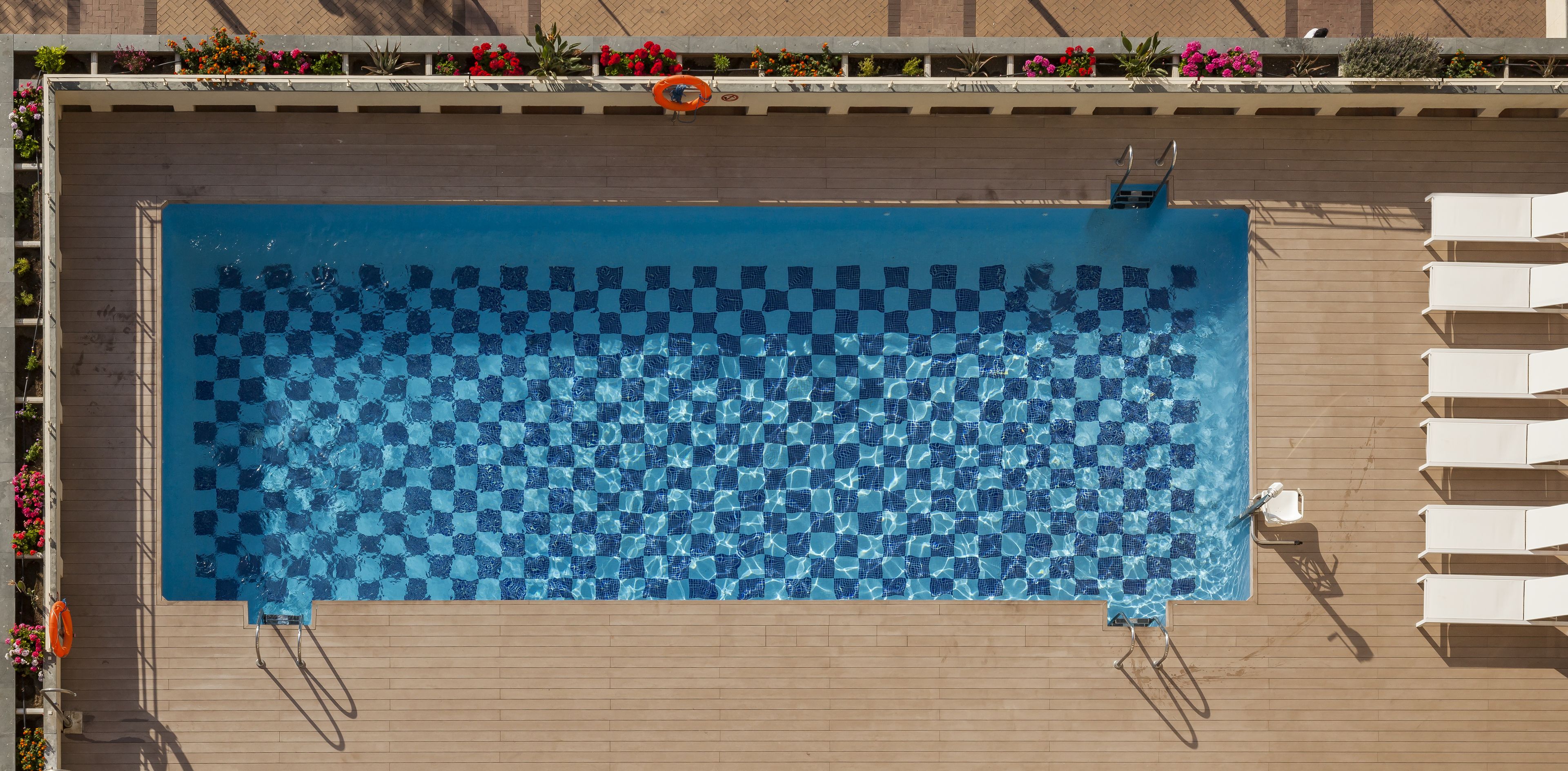 Una piscina rectangular amb un mosaic de rajoles de disseny dairellat, envoltada per un terra de fusta i gandules blanques.