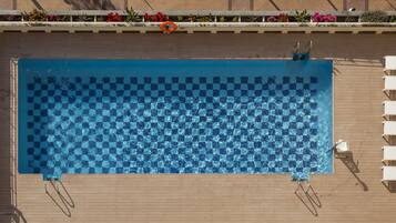 Una piscina rectangular amb un mosaic de rajoles de disseny dairellat, envoltada per un terra de fusta i gandules blanques.