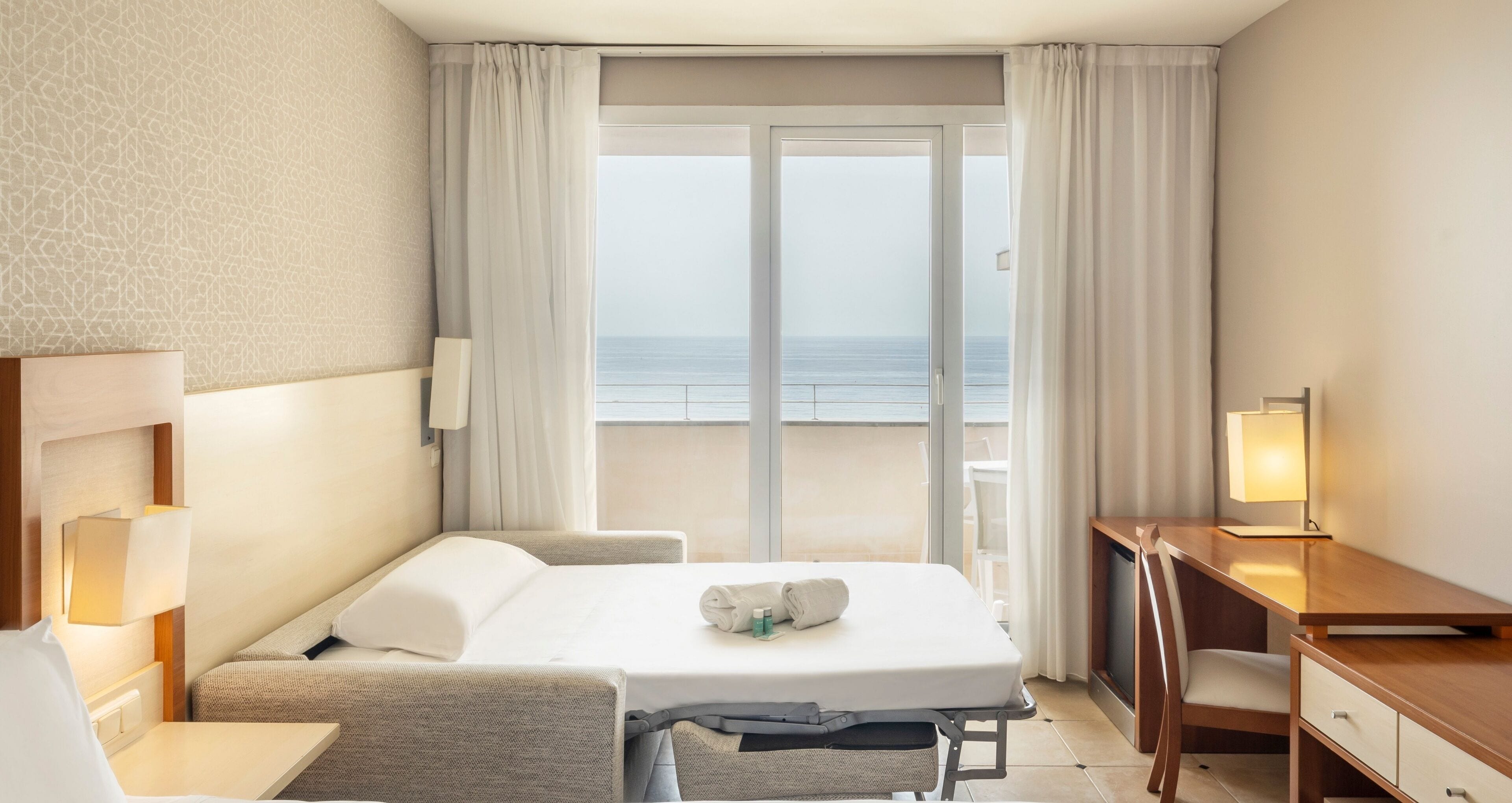 A hotel room with a bed, a desk, a chair, a lamp, and a view of the sea through a large window.