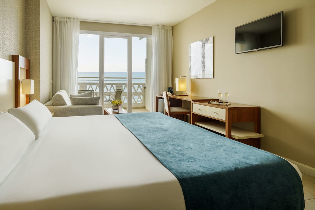 A hotel room with a large bed, a desk, a chair, a TV, and a view of the sea.