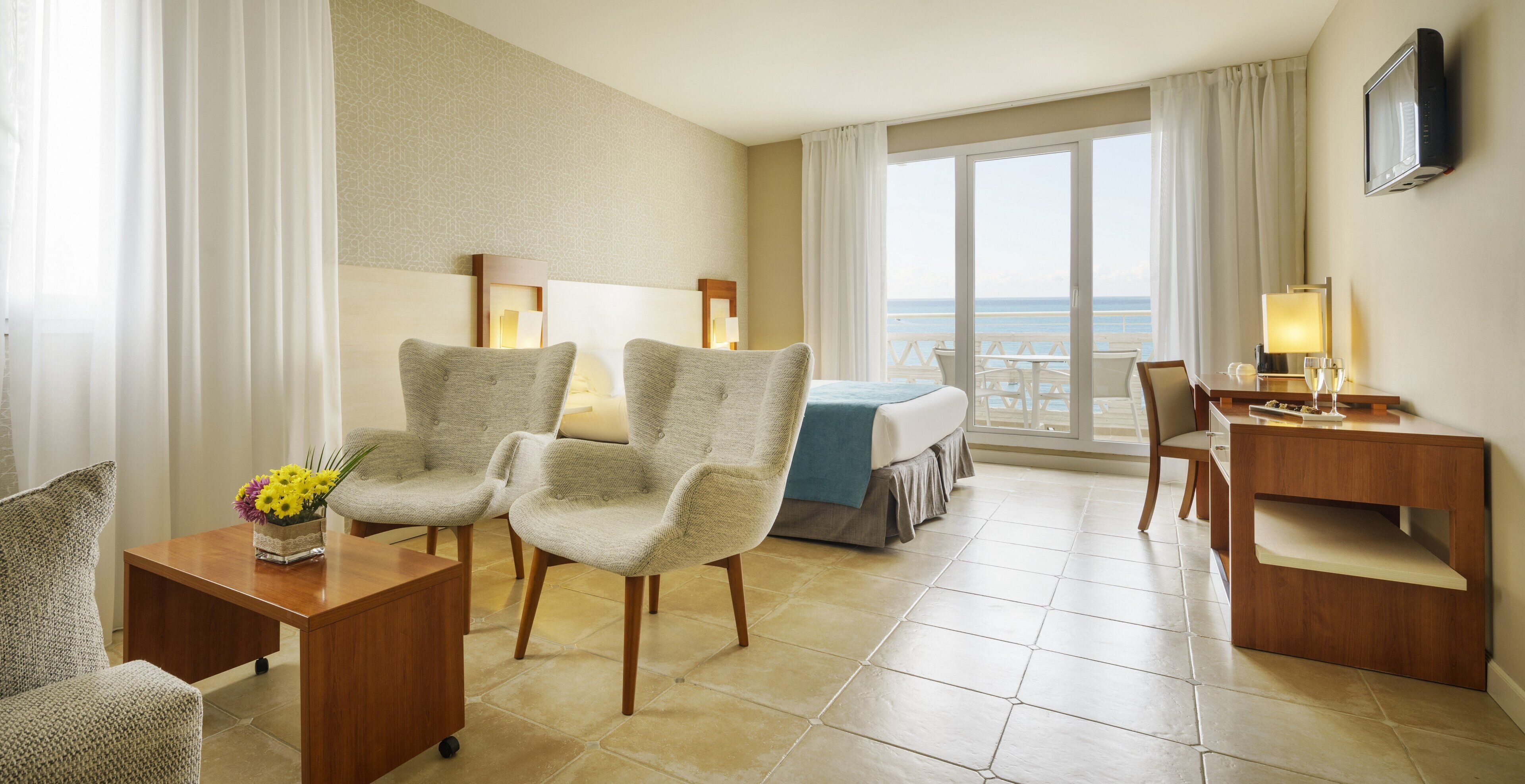 A hotel room with a bed, two armchairs, a desk, and a view of the sea.
