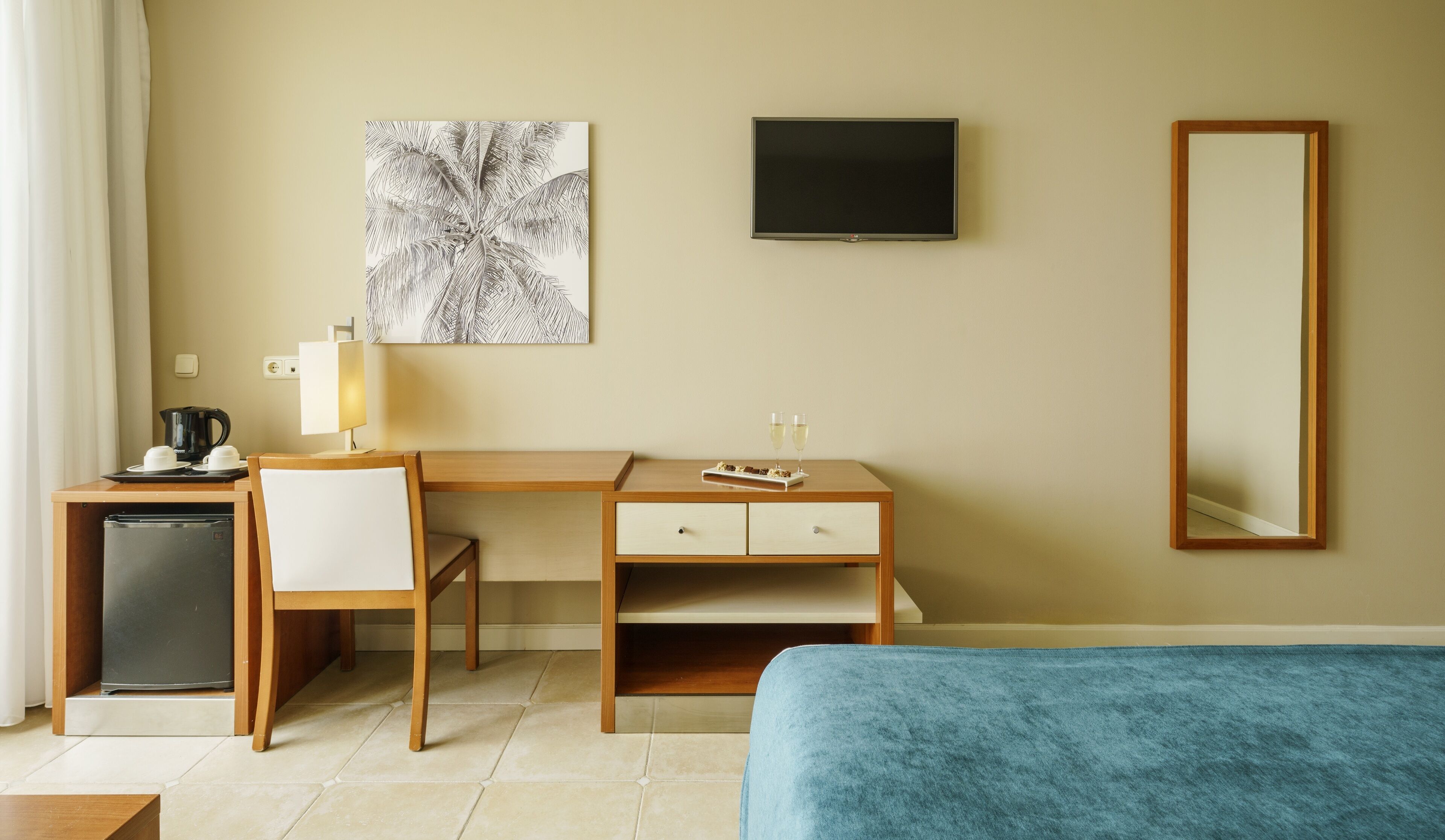 A hotel room with a desk, chair, small refrigerator, bedside table, and a bed.