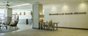 Un área de recepción moderna con sillas de mimbre, una mesa de madera y una pared con un mensaje de bienvenida.