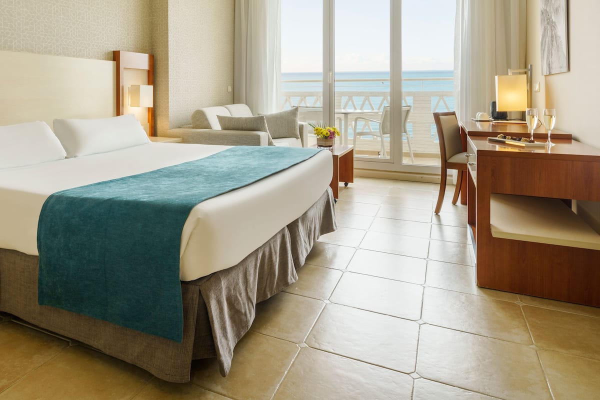 A hotel room with a large bed, a desk, a chair, and a view of the sea.