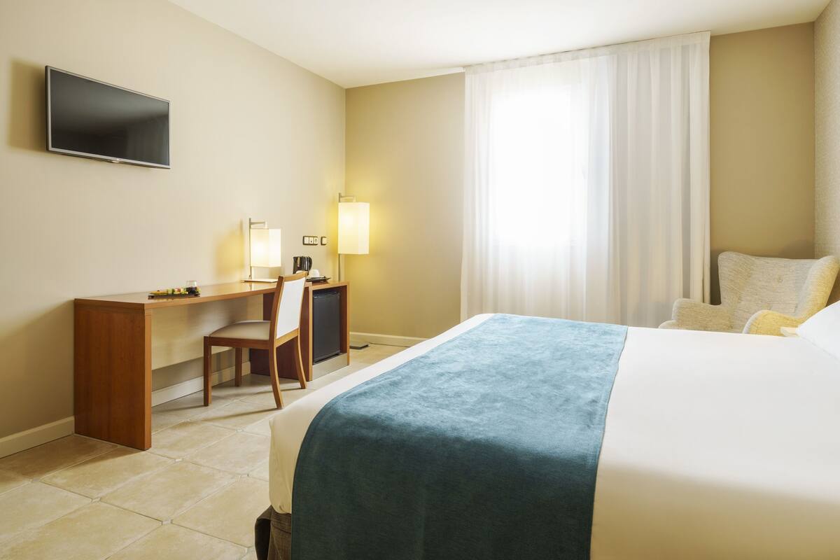 A hotel room with a bed, desk, chair, TV, and a window with curtains.