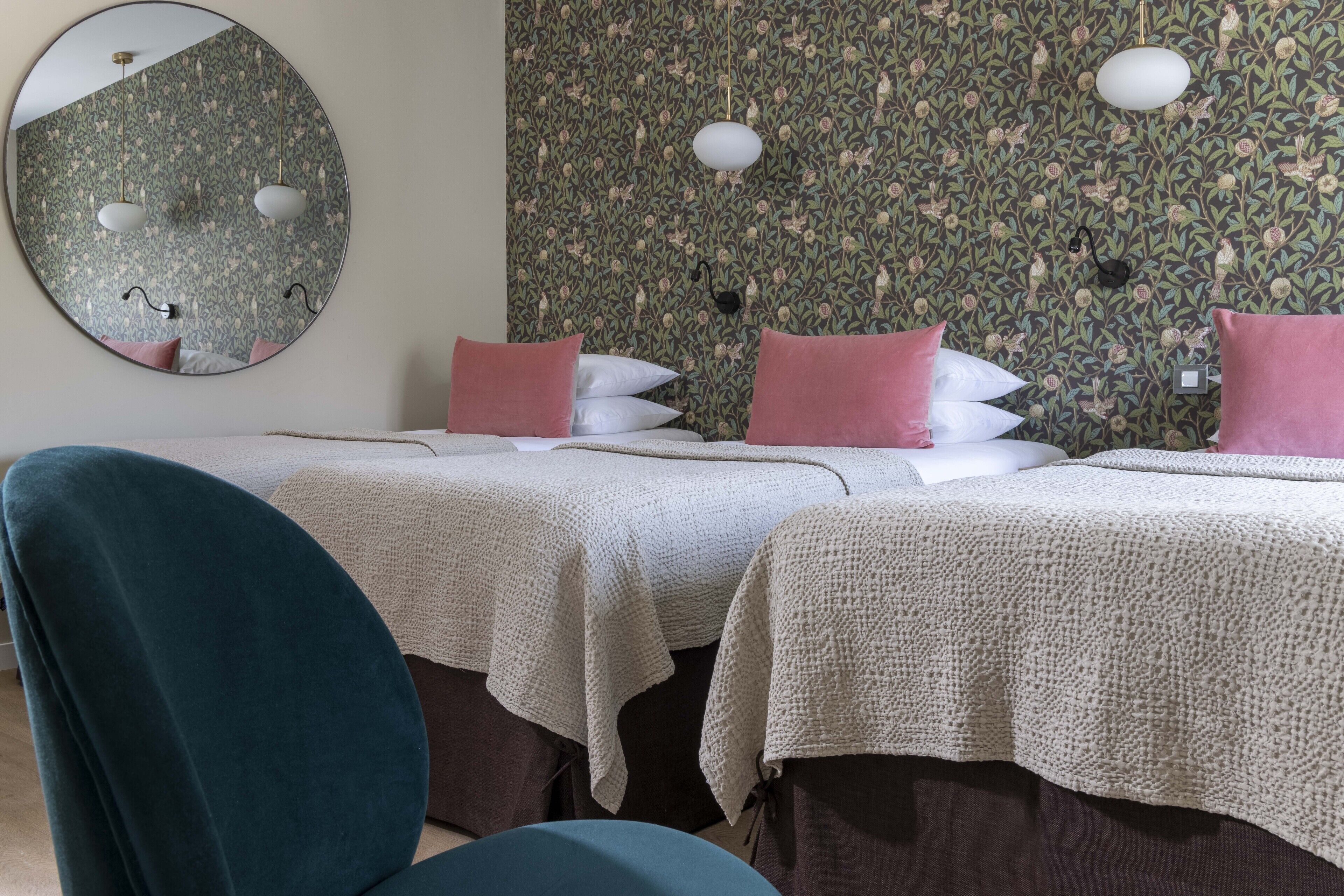A hotel room with two beds, a round mirror, floral wallpaper, and a teal chair.