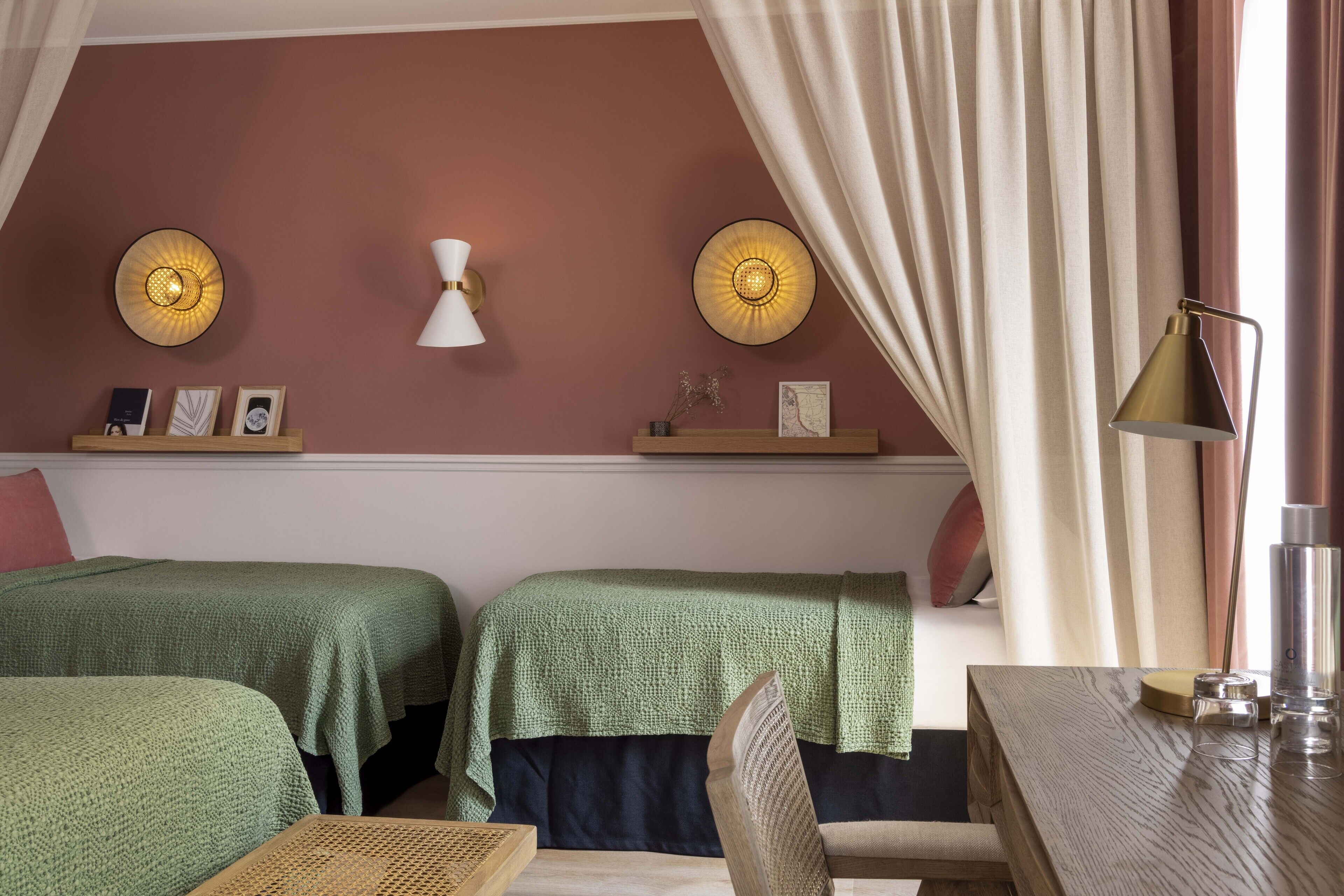 Une chambre d’hôtel avec deux lits verts, une table en bois, une lampe et un luminaire fixé au mur.