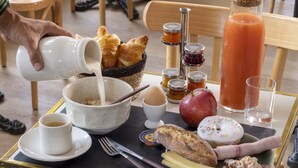 Bandeja de desayuno con cruasanes, pan, queso, jamón, una rosquilla, una manzana, un vaso de jugo, una taza de café y un tazón de cereal.