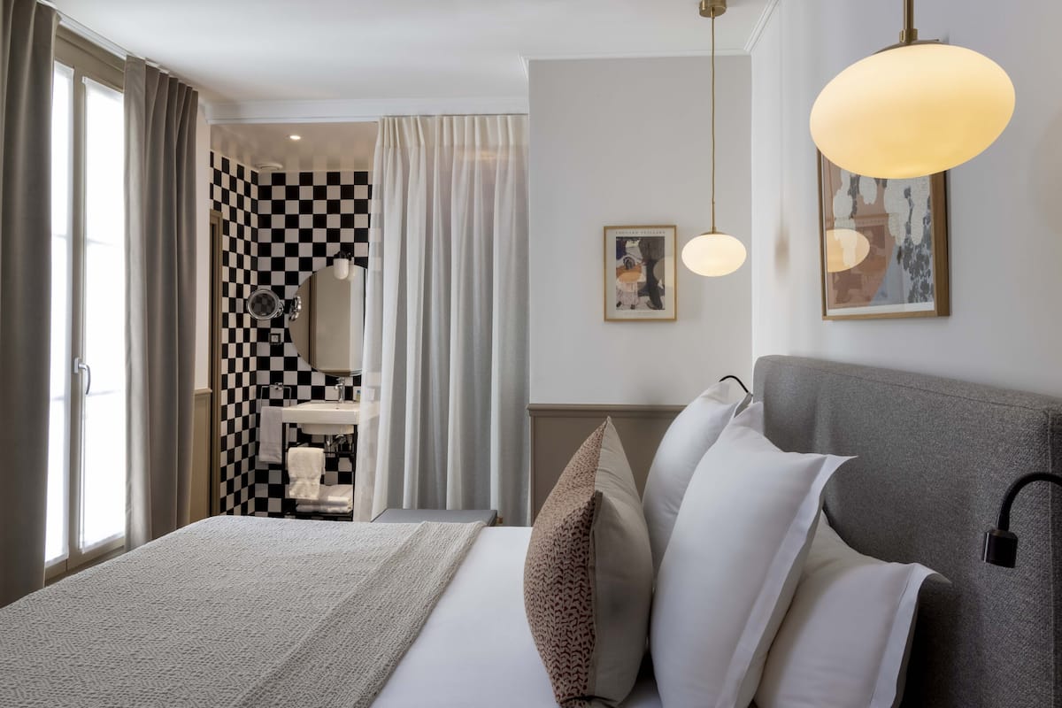 Ein Hotelzimmer mit karierter Wand, einem Bett mit vielen Kissen, einem Nachttisch, einem Spiegel und einer Pendelleuchte.